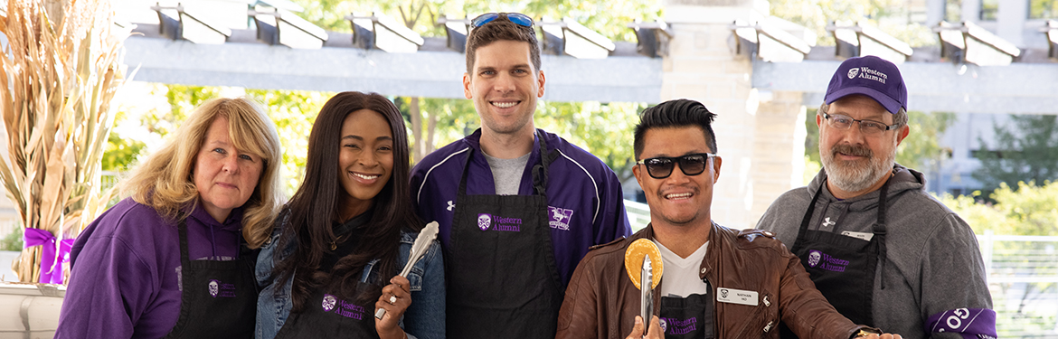 A group of alumni smile behind a BBQ