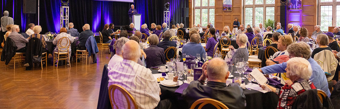 Alumni sit at tables and listen to speaker at a podium