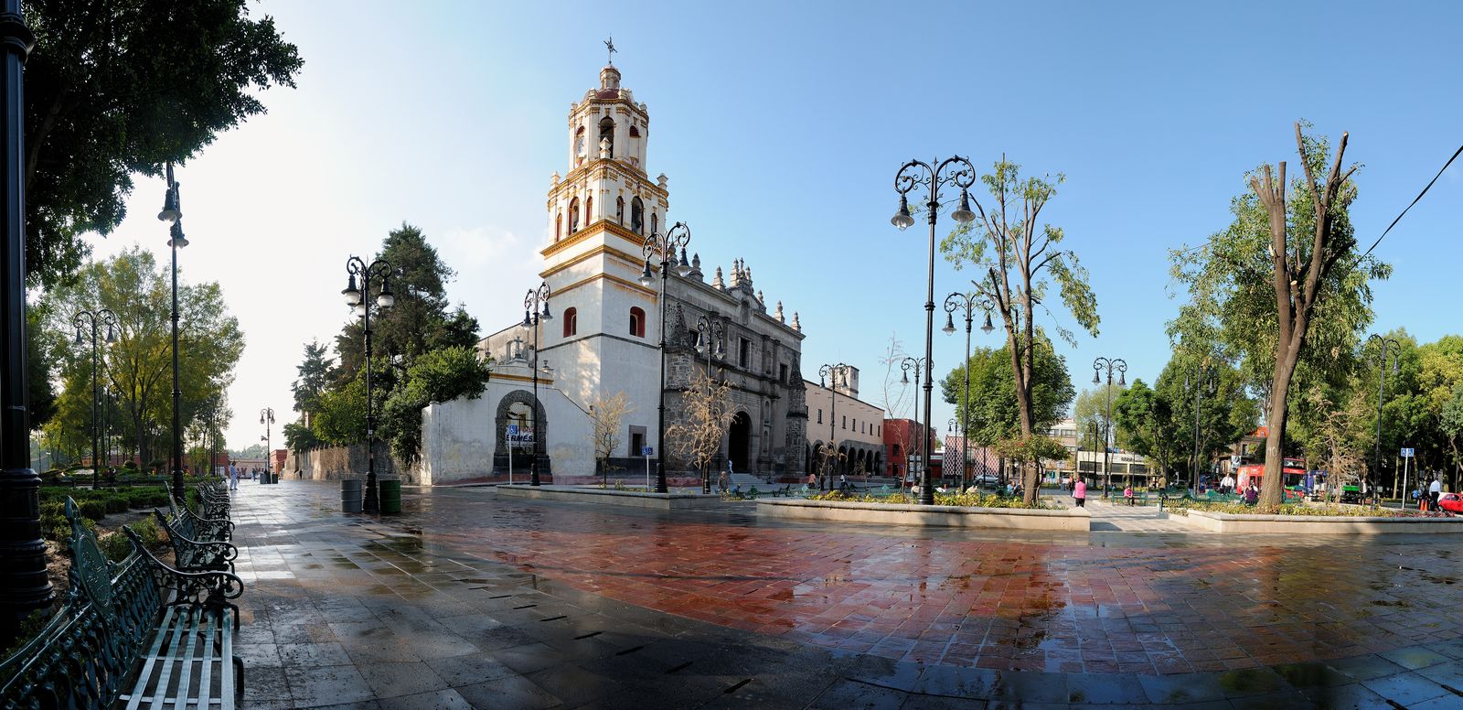 Historic bell-towered church, wet Coyoacán plaza, green trees, and pedestrians under clear blue sky